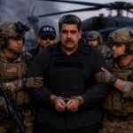 Nicolas Maduro in handcuffs escorted by U.S. military personnel during a detention operation, with a military helicopter in the background.