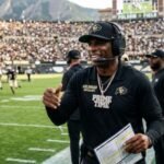 "Deion Sanders" Sanders, wearing a black headset, CU cap, and a "PRIME TIME" diamond necklace