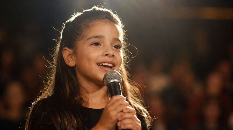 Young girl singing confidently into a microphone on a softly lit stage with a blurred audience in the background, symbolizing early talent and rising fame.