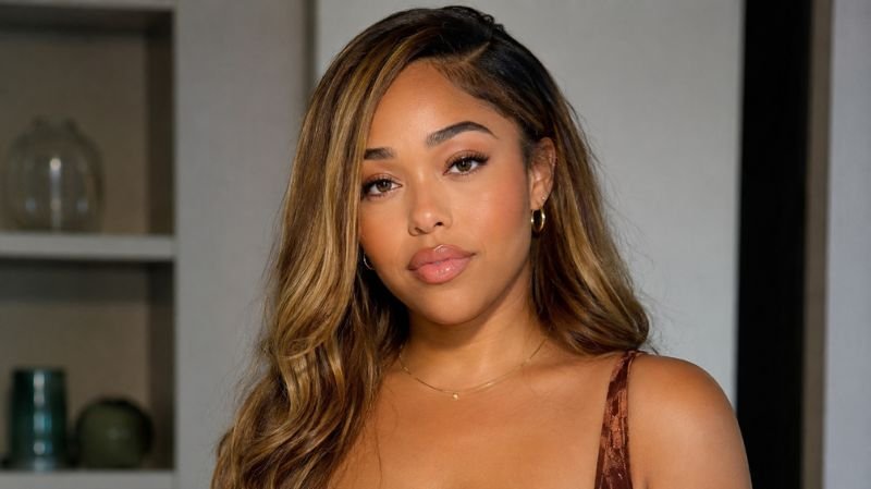 Portrait of a stylish young woman with highlighted wavy hair, wearing subtle jewelry and neutral-toned clothing, posing indoors with soft lighting and a blurred modern background.