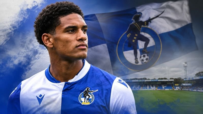 A dramatic portrait of a young footballer in a blue and white kit standing in front of a stadium backdrop with club flag imagery and smoky effects, representing a loan move to Bristol Rovers.